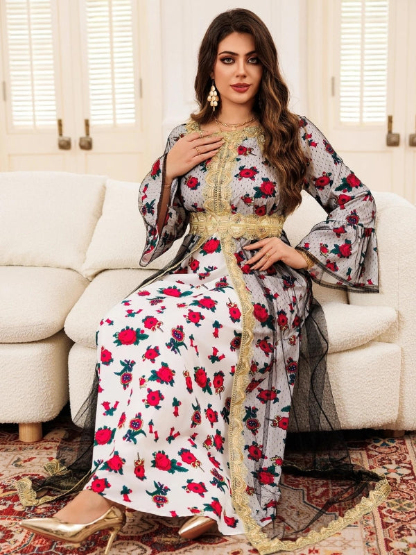 La femme porte un magnifique caftan long à motif floral rouge et vert sur un fond gris, orné de bordures dorées qui ajoutent une touche d'élégance au vêtement. Le caftan est ajusté à la taille avec une ceinture dorée qui souligne sa silhouette. Les manches évasées apportent une touche de fluidité et de grâce à l'ensemble. Ses cheveux longs et ondulés tombent élégamment sur ses épaules. Elle se tient debout dans un intérieur chaleureux, devant un canapé clair, avec un tapis à motifs sous ses pieds