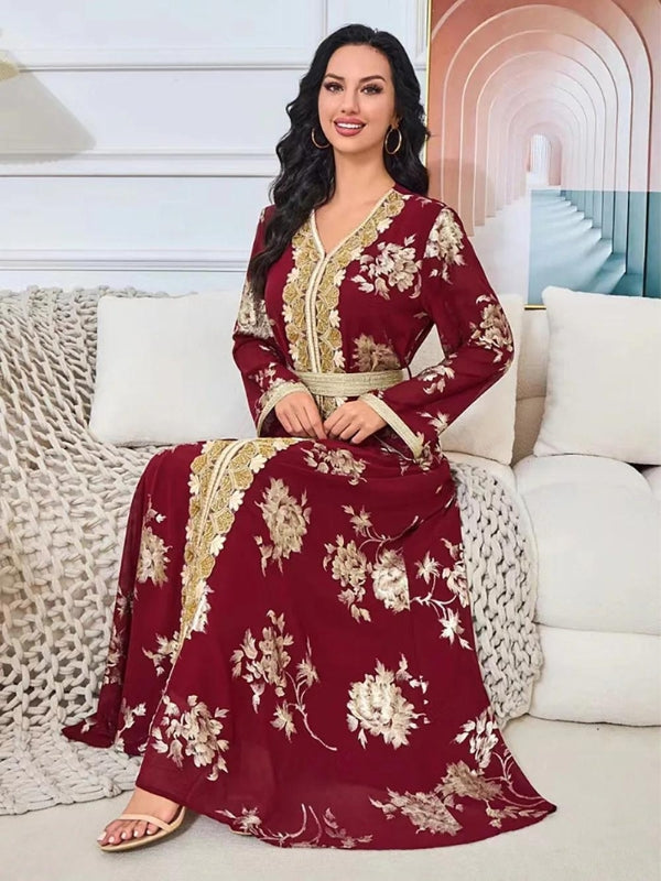 La femme est assise, souriante, portant un caftan bordeaux avec des motifs floraux dorés. Le caftan est richement décoré avec une bordure dorée le long du devant, et une ceinture dorée souligne sa taille. Ses cheveux noirs et ondulés tombent librement sur ses épaules. Elle porte également des boucles d'oreilles dorées qui complètent son look. Elle est assise sur un canapé clair, avec un plaid texturé à côté d'elle, dans une pièce au décor moderne et élégant, comprenant un mur avec des arches en arrière-plan