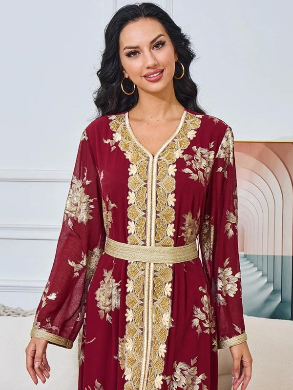 La femme est assise, souriante, portant un caftan bordeaux avec des motifs floraux dorés. Le caftan est richement décoré avec une bordure dorée le long du devant, et une ceinture dorée souligne sa taille. Ses cheveux noirs et ondulés tombent librement sur ses épaules. Elle porte également des boucles d'oreilles dorées qui complètent son look. Elle est assise sur un canapé clair, avec un plaid texturé à côté d'elle, dans une pièce au décor moderne et élégant, comprenant un mur avec des arches en arrière-plan