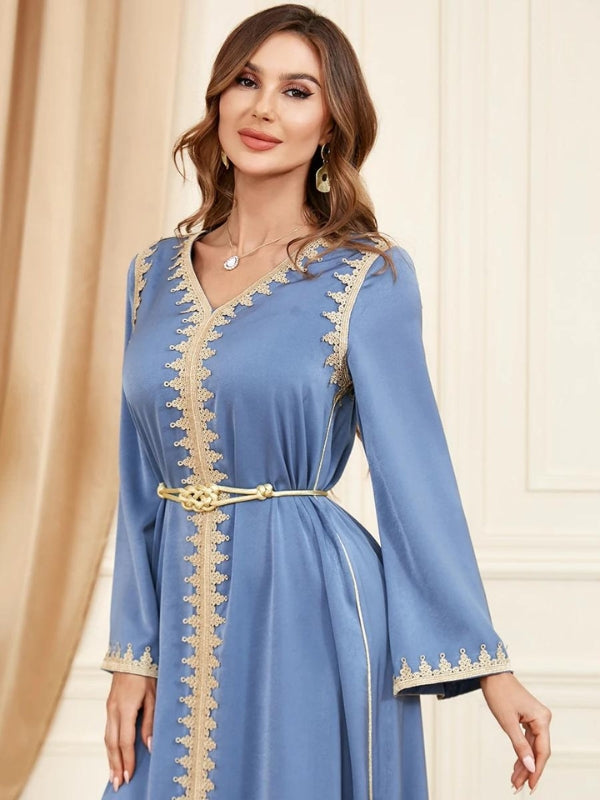 La femme est vêtue d'un élégant caftan bleu clair, qui présente une coupe longue et fluide, accentuée par des détails dorés le long du bord avant et des manches. Le caftan est resserré à la taille par une ceinture dorée, ajoutant une touche de structure à la tenue. Les manches évasées et le décolleté en V orné de broderies dorées soulignent la finesse et la sophistication du design