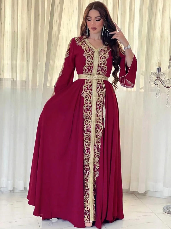 Une femme portant un somptueux caftan bordeaux richement orné de broderies dorées qui descendent le long du centre de la robe et sur les manches. Le caftan, qui épouse gracieusement la silhouette, est cintré à la taille par une ceinture assortie, également ornée de détails dorés. Le tissu fluide et ample confère à la tenue une allure majestueuse, tandis que la couleur bordeaux apporte une touche de raffinement et de chaleur. L'arrière-plan, composé de rideaux légers, met en valeur la tenue