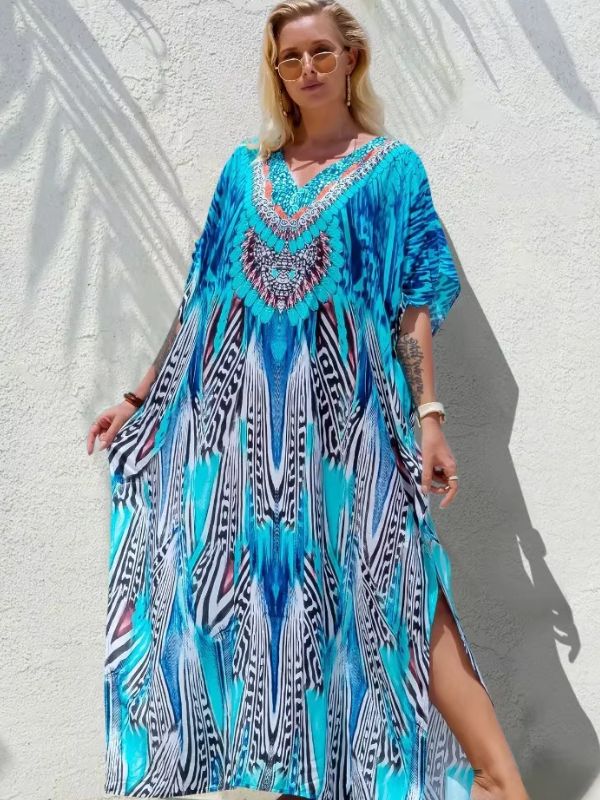 Une femme habillée d’une djellaba fluide turquoise aux motifs inspirés des plumes, posant contre un mur clair