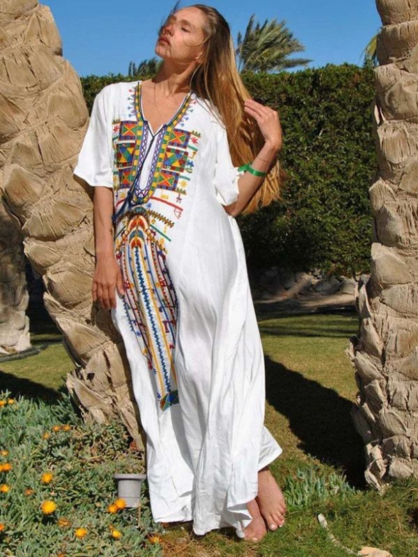 Femme debout entre deux palmiers portant une djellaba blanche brodée de motifs berbères multicolores