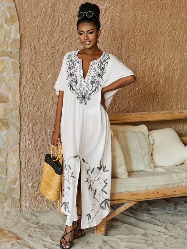 Femme debout près d’un mur en pierre, vêtue d’une djellaba blanche à col brodé et sac en osier à la main