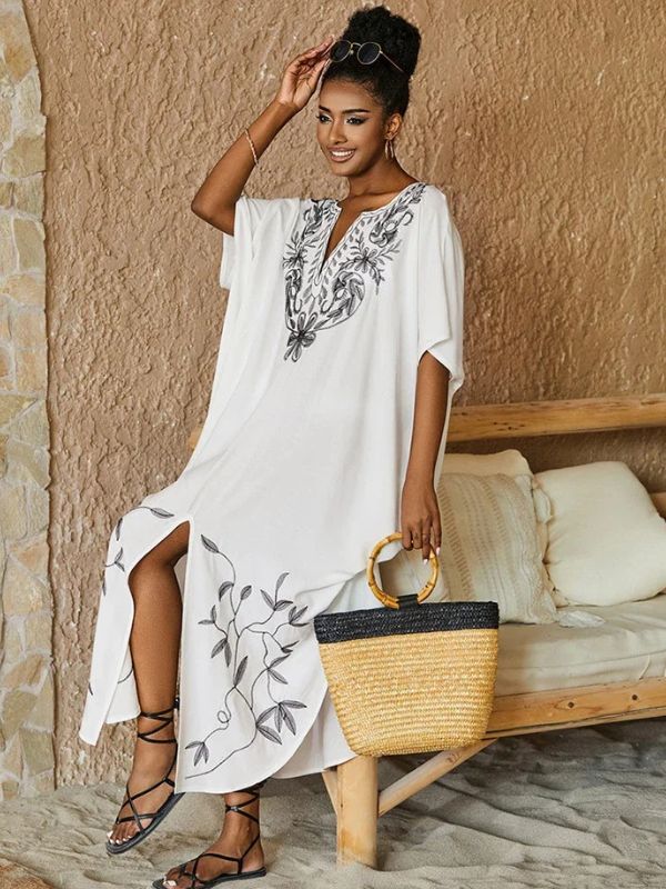 Femme debout près d’un mur en pierre, vêtue d’une djellaba blanche à col brodé et sac en osier à la main