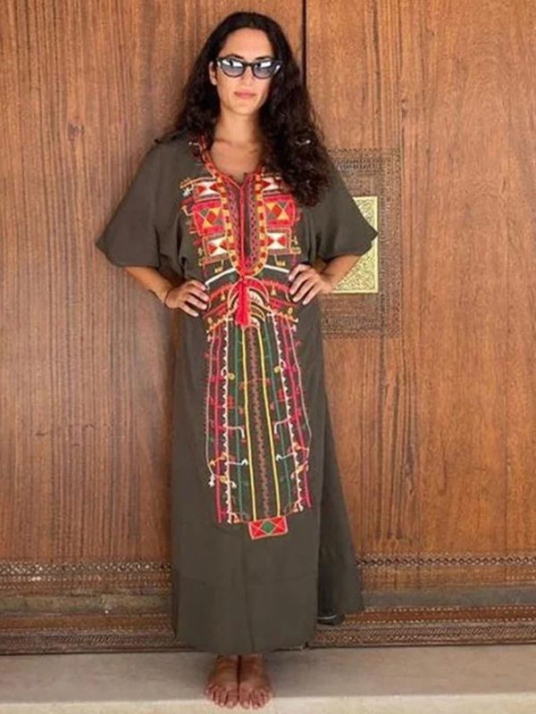 Une femme porte une djellaba kaki à broderies amazighes rouges et jaunes, posant debout les mains sur les hanches