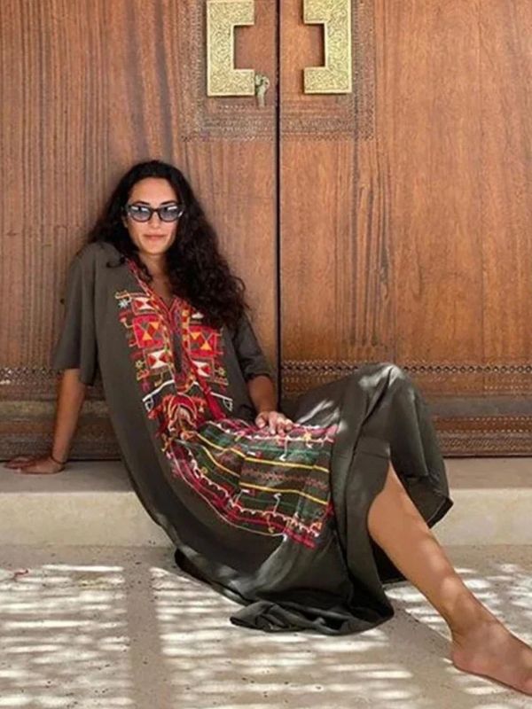 Une femme porte une djellaba kaki à broderies amazighes rouges et jaunes, posant debout les mains sur les hanches