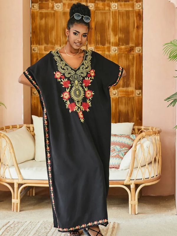 Femme debout en djellaba noire décorée de broderies dorées et rouges, posant devant un décor intérieur en rotin