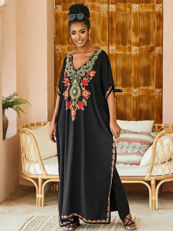 Femme souriante en djellaba noire à motifs floraux rouges et dorés, posant dans un salon au style bohème