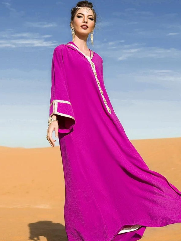 Femme debout dans le désert portant une djellaba marocaine fuchsia fluide avec des finitions dorées, posant sous un ciel bleu clair