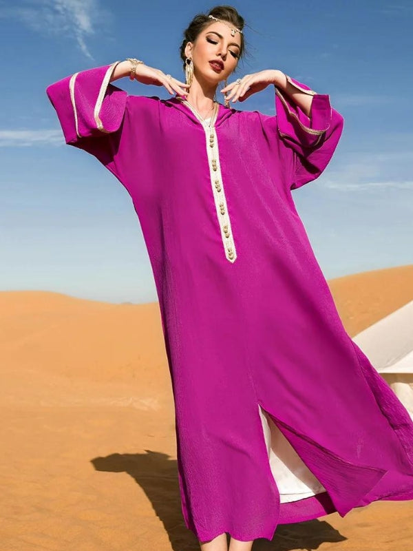 Femme en djellaba marocaine fuchsia avec des détails dorés, les bras levés, posant dans un désert sous un ciel bleu