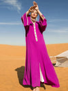 Femme en djellaba marocaine fuchsia avec des bordures dorées, levant les bras face au soleil dans le désert sous un ciel bleu dégagé