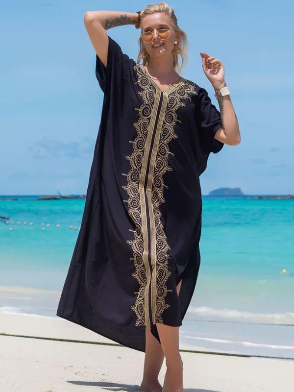 Femme en djellaba noire avec broderies dorées, levant la main en souriant sur une plage tropicale