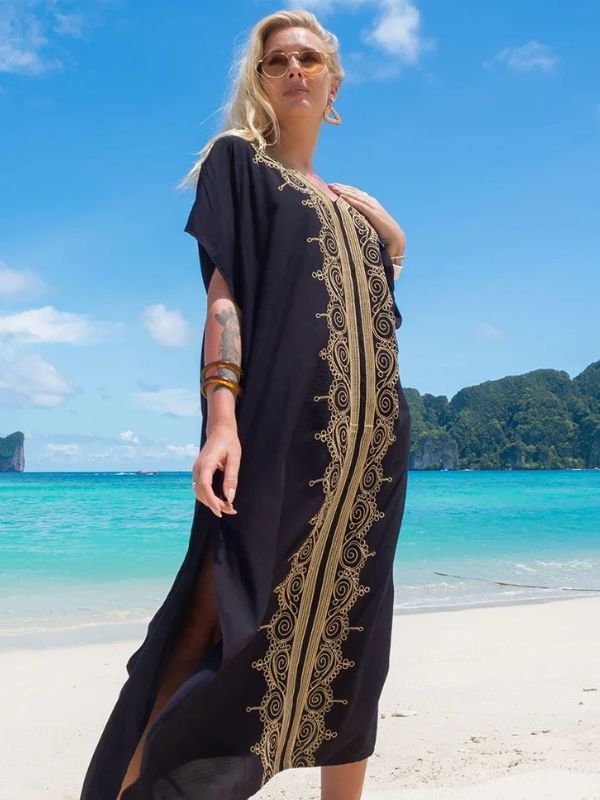 Femme en djellaba noire avec broderies dorées verticales, posant sur une plage avec vue sur des montagnes verdoyantes