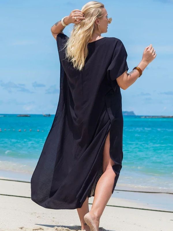 Femme de dos portant une djellaba noire fluide, marchant sur une plage au bord d’une mer turquoise