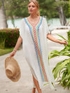 Femme élégante en djellaba blanche avec décorations colorées, marchant sur une plage tropicale avec un chapeau à la main