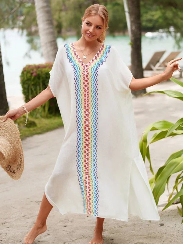 Femme tenant un chapeau de paille et portant une djellaba blanche décorée de motifs multicolores, se promenant sur une plage