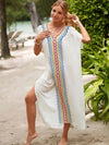 Femme portant une djellaba blanche ornée de motifs zigzag multicolores, marchant pieds nus sur une plage tropicale