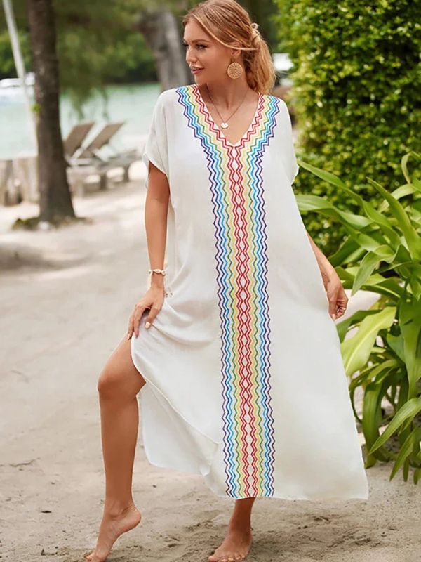 Femme souriante en djellaba blanche à motifs zigzag arc-en-ciel, posant sur une plage au bord de l’eau