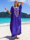 Femme portant une djellaba bleu roi à motifs dorés, posant sur une plage avec vue sur des montagnes verdoyantes