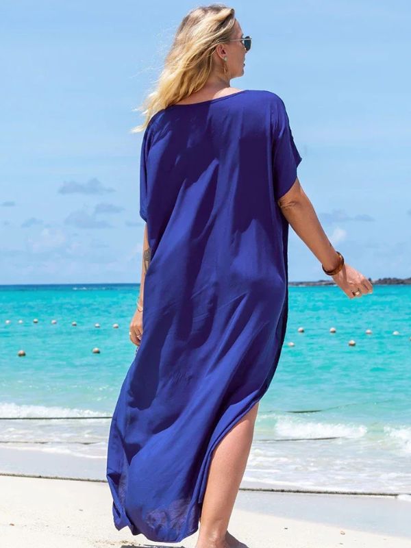 Femme de dos portant une djellaba longue bleu roi, marchant pieds nus sur une plage au bord d’une mer turquoise