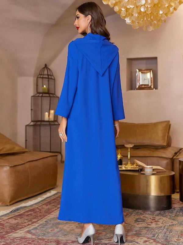 La femme est vue de dos, portant une élégante djellaba bleu vif à capuche qui descend jusqu&#39;aux chevilles. La coupe ample de la djellaba crée une silhouette fluide, tandis que la capuche repose délicatement sur ses épaules. Elle complète son look avec des escarpins argentés à talons, ajoutant une touche de sophistication. Ses cheveux sont lâchés, tombant légèrement sur son dos