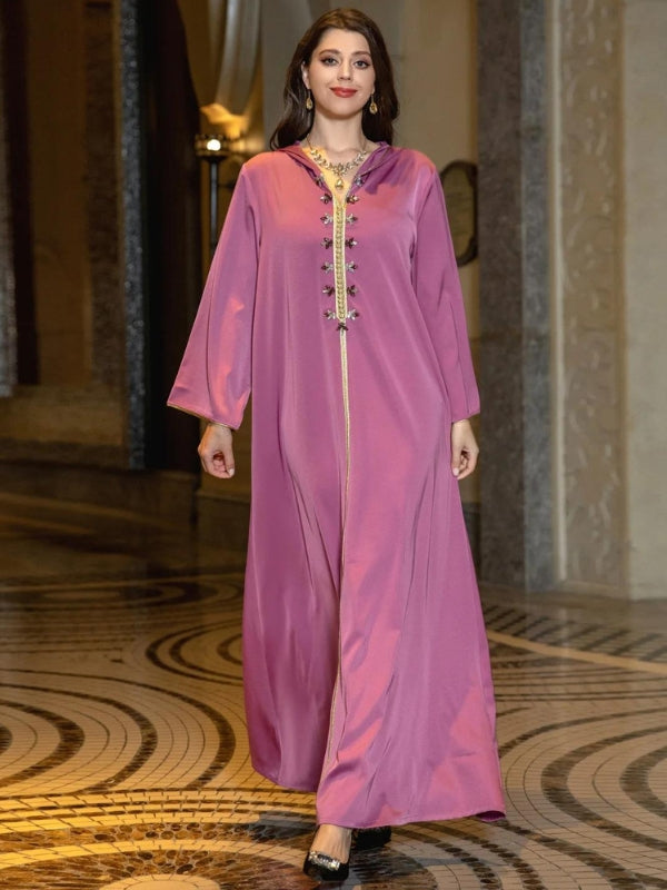 Une femme portant une djellaba rose sable avec des ornements dorés le long du col et de la fermeture avant, se tient avec assurance. Ses cheveux sont détachés, et elle complète son look avec des boucles d'oreilles dorées et un collier assorti