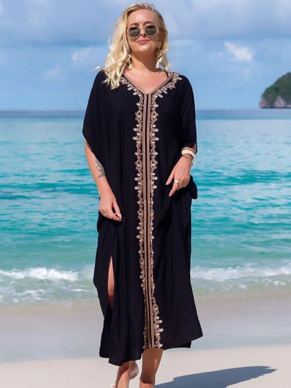 Femme debout sur le sable, portant une djellaba noire élégante à décorations dorées, face à la mer turquoise