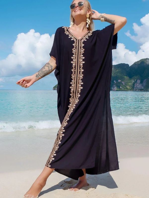 Femme souriante en djellaba noire à broderie dorée, levant légèrement le bras sur une plage exotique