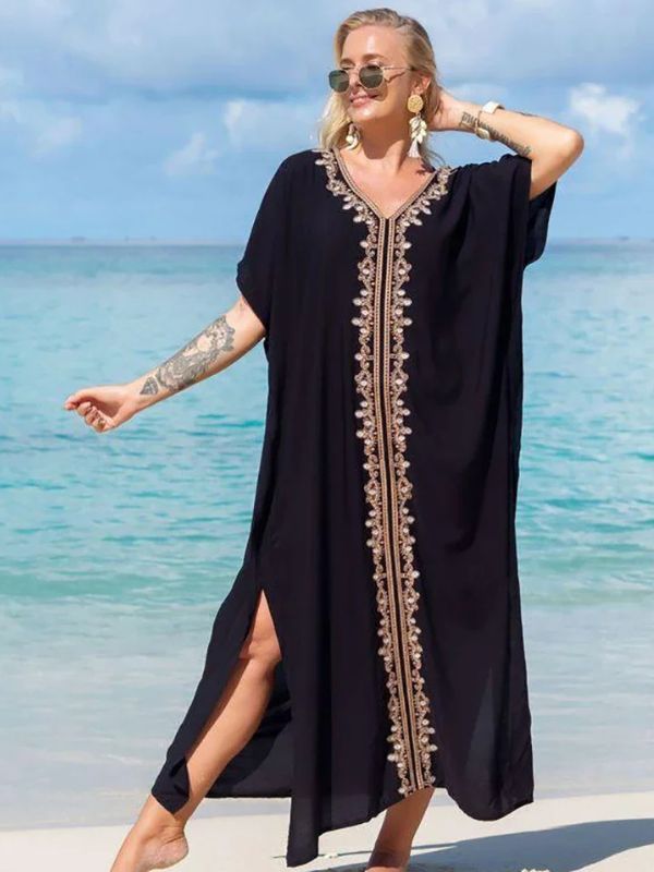 Femme souriante en djellaba noire décorée d’une broderie dorée verticale, posant sur une plage tropicale