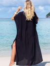 Femme de dos portant une djellaba noire fluide, marchant sur une plage au bord d’une mer turquoise