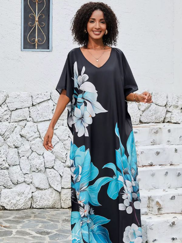 Une femme porte une djellaba noire ornée de grands motifs floraux bleus et blancs, posant devant un mur en pierre et des escaliers blancs