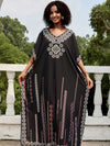 Une femme porte une djellaba noire ornée de broderies géométriques colorées, posant sur une terrasse blanche avec une balustrade et des arbres en arrière-plan