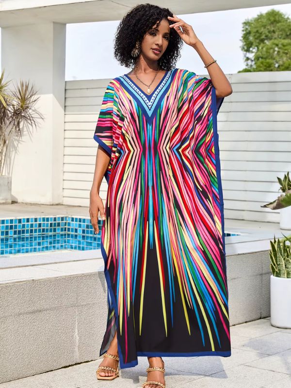 Une femme porte une djellaba noire ornée de rayures colorées vives, posant près d’une piscine avec des plantes autour.