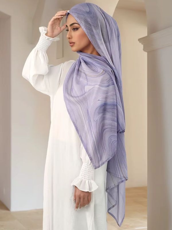 Hijab en coton imprimé lavande, porté, main relevée vers le front, lignes fines en volutes sur fond lilas