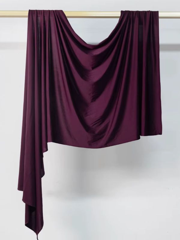 Hijab en jersey violet foncé aubergine suspendu et drapé sur une tringle dorée, présenté sur fond blanc