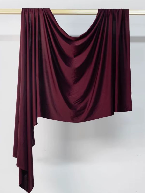 Hijab en jersey bordeaux profond suspendu et drapé sur une tringle dorée, présenté sur fond blanc
