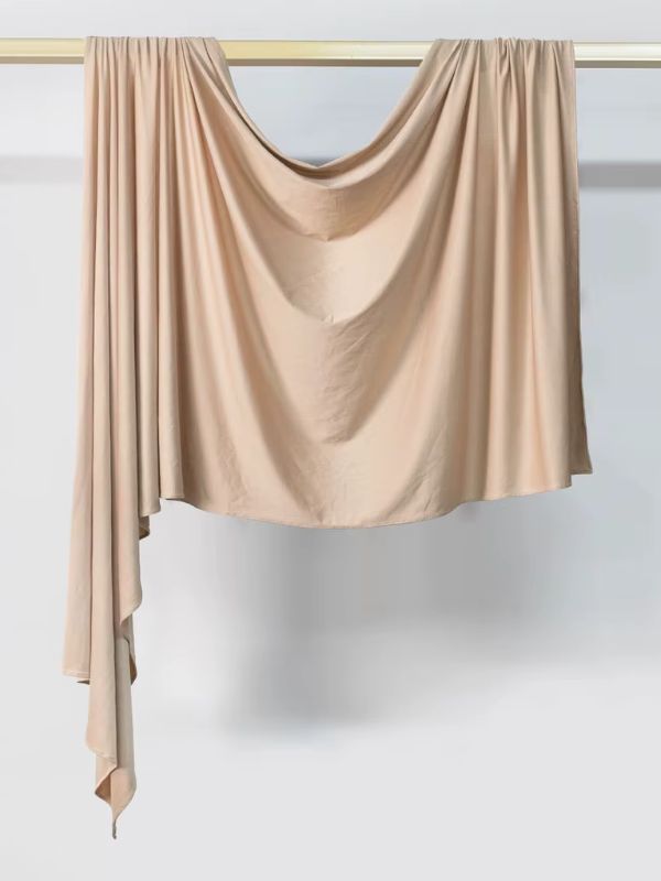 Hijab en jersey beige sable suspendu et drapé sur une tringle dorée, présenté sur fond blanc