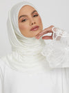 Une femme porte un hijab blanc, posé élégamment, avec une main délicatement levée vers son visage. La manche de sa tenue est ornée de dentelle florale, ajoutant une touche raffinée. Son regard est doux et confiant