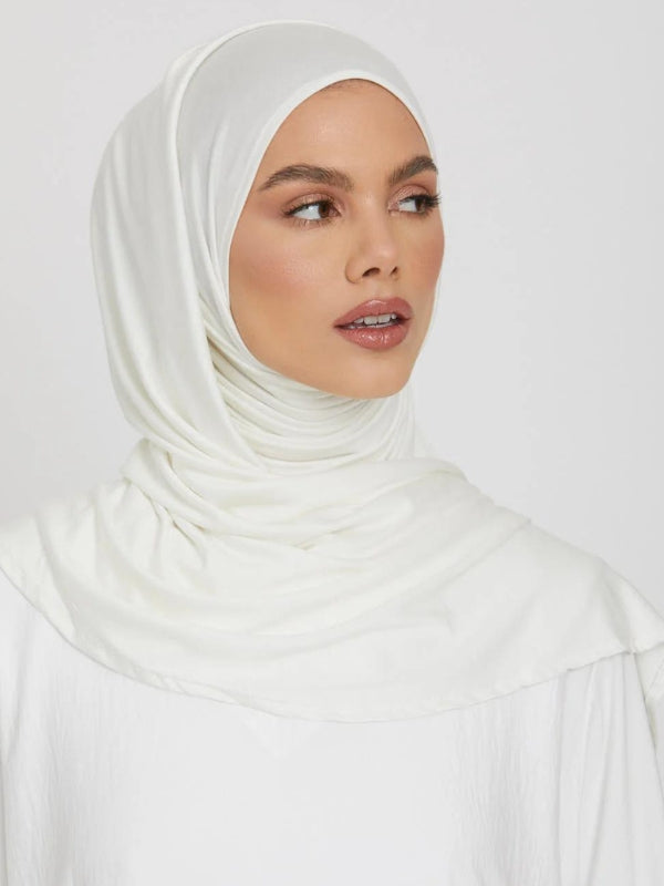 Une femme porte un hijab blanc, posé élégamment, avec une main délicatement levée vers son visage. La manche de sa tenue est ornée de dentelle florale, ajoutant une touche raffinée. Son regard est doux et confiant