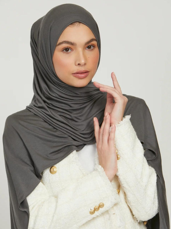 Une femme porte un hijab en jersey gris, qui entoure élégamment son visage. Elle est habillée d'une veste blanche avec des boutons dorés, ajoutant une touche de sophistication à son look