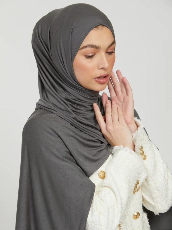 Une femme porte un hijab en jersey gris, qui entoure élégamment son visage. Elle est habillée d'une veste blanche avec des boutons dorés, ajoutant une touche de sophistication à son look