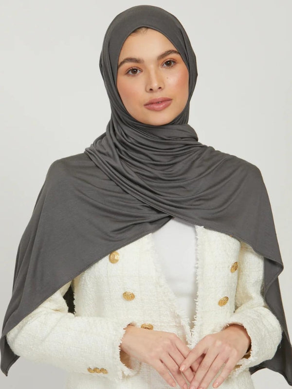 Une femme porte un hijab en jersey gris, couvrant ses cheveux et ses épaules. Elle est vêtue d&#39;une veste blanche texturée avec des boutons dorés, ses mains jointes devant elle, adoptant une posture calme et assurée