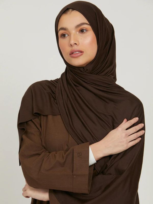 Femme portant un hijab en jersey marron foncé, posant avec une main sur son bras, le regard vers l'avant