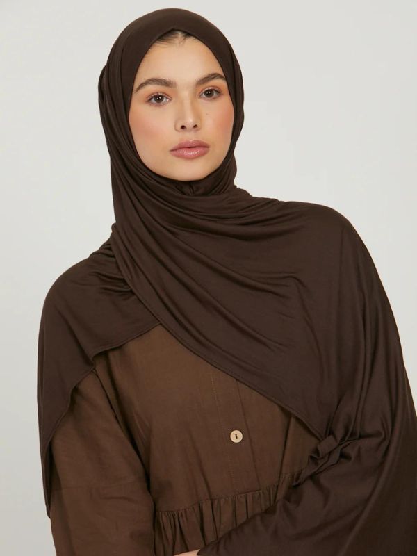 Femme portant un hijab en jersey marron foncé, posant avec une main sur son bras, le regard vers l'avant