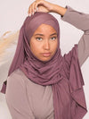 Une femme portant un hijab de couleur mauve. Le hijab est drapé de manière élégante autour de sa tête et de son cou, avec un style qui recouvre bien le front et tombe doucement autour de son épaule. La femme a une expression douce et sereine. Le tissu du hijab semble être en matière légère et confortable, et le drapé met en valeur les contours de son visage. L&#39;ensemble est harmonieux avec la tenue de couleur similaire qu&#39;elle porte, créant un look coordonné et raffiné