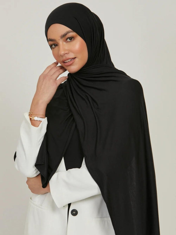 La photo montre une femme portant un hijab noir, drapé élégamment autour de sa tête et tombant sur ses épaules. Elle porte également un manteau blanc, créant un contraste sophistiqué avec le hijab. La femme a un léger sourire, et sa main droite est levée près de son visage, ajoutant une touche de douceur et de grâce à sa posture. Le hijab est bien ajusté et semble être fait d'un tissu léger et fluide, accentuant l'élégance de l'ensemble