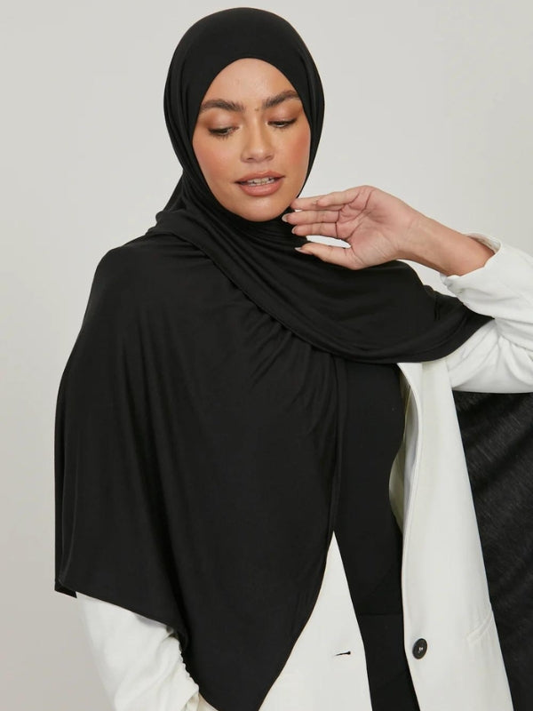 La photo montre une femme portant un hijab noir, drapé élégamment autour de sa tête et tombant sur ses épaules. Elle porte également un manteau blanc, créant un contraste sophistiqué avec le hijab. La femme a un léger sourire, et sa main droite est levée près de son visage, ajoutant une touche de douceur et de grâce à sa posture. Le hijab est bien ajusté et semble être fait d'un tissu léger et fluide, accentuant l'élégance de l'ensemble