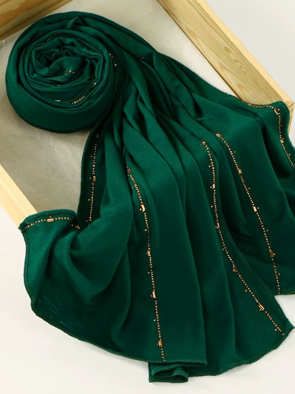 Hijab jersey premium strassé vert sapin, enroulé et drapé dans un cadre en bois, lignes de strass cuivrés et dorés sur toute la longueur