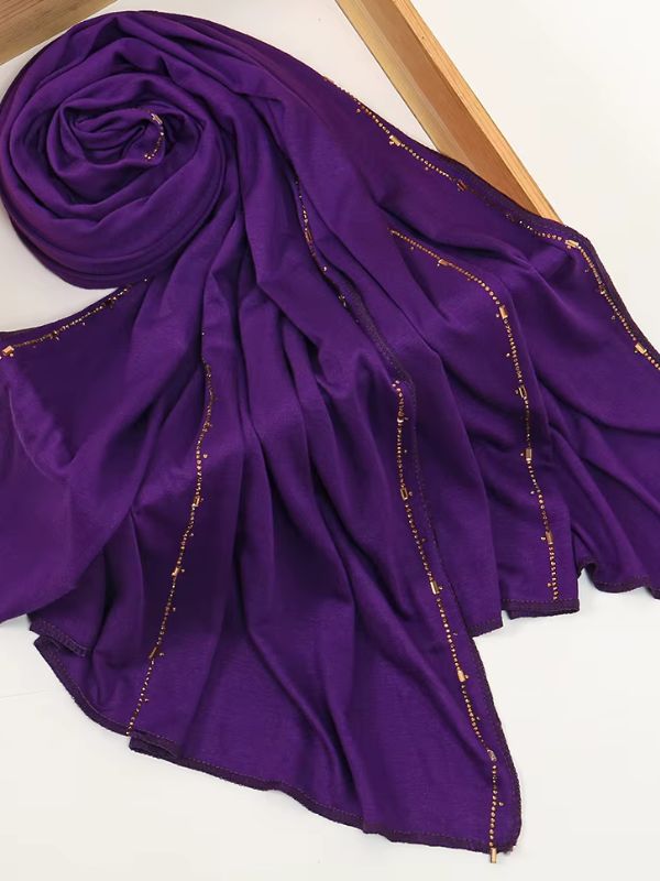 Hijab jersey premium strassé violet profond, enroulé et drapé dans un cadre en bois, lignes de strass dorés sur toute la longueur